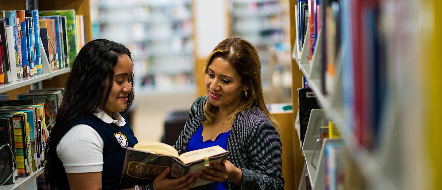 一个成年人和一个学生在图书馆的满书架之间拿着一本打开的书.